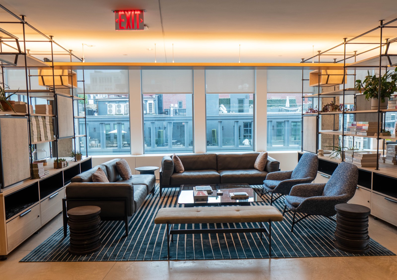 Two leather sofas and a pair of upholstered chairs on a striped rug, between blackened steel and oak shelving that runs floor to ceiling on both sides, with full-height glazing and roller shades behind
