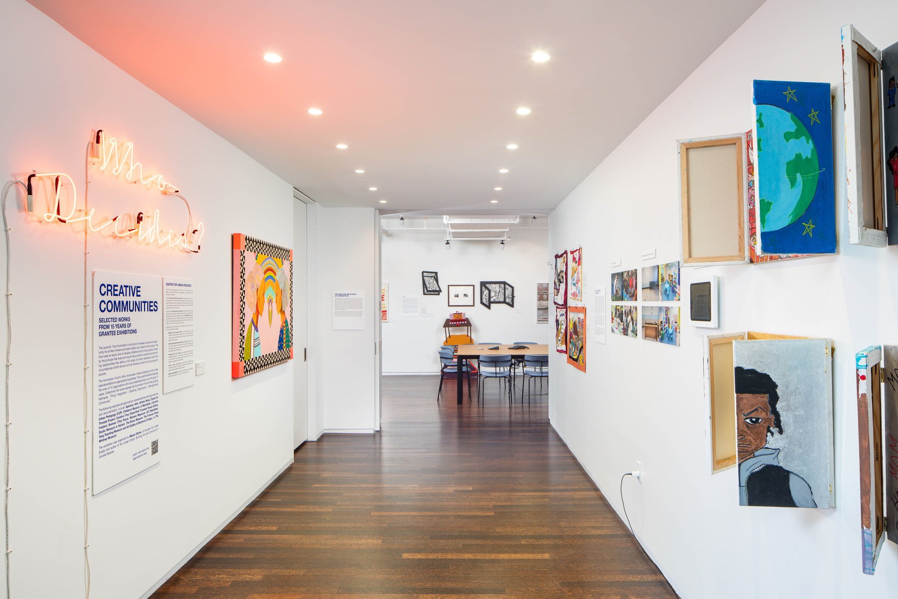 A corridor hung with framed and unframed work along both walls, a neon script piece and an exhibition placard at the near end, a run of recessed downlights overhead, and a meeting table at the far end