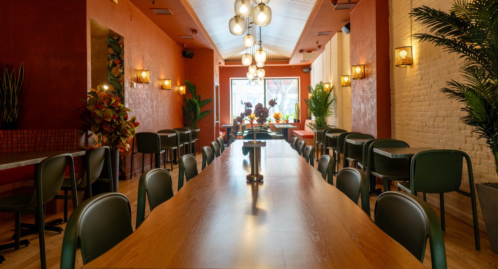 A long oak communal table down the center of the room with dark green chairs either side, under a cluster of glass globe pendants and a ribbed barrel ceiling, between terracotta plaster on one wall and painted white brick on the other