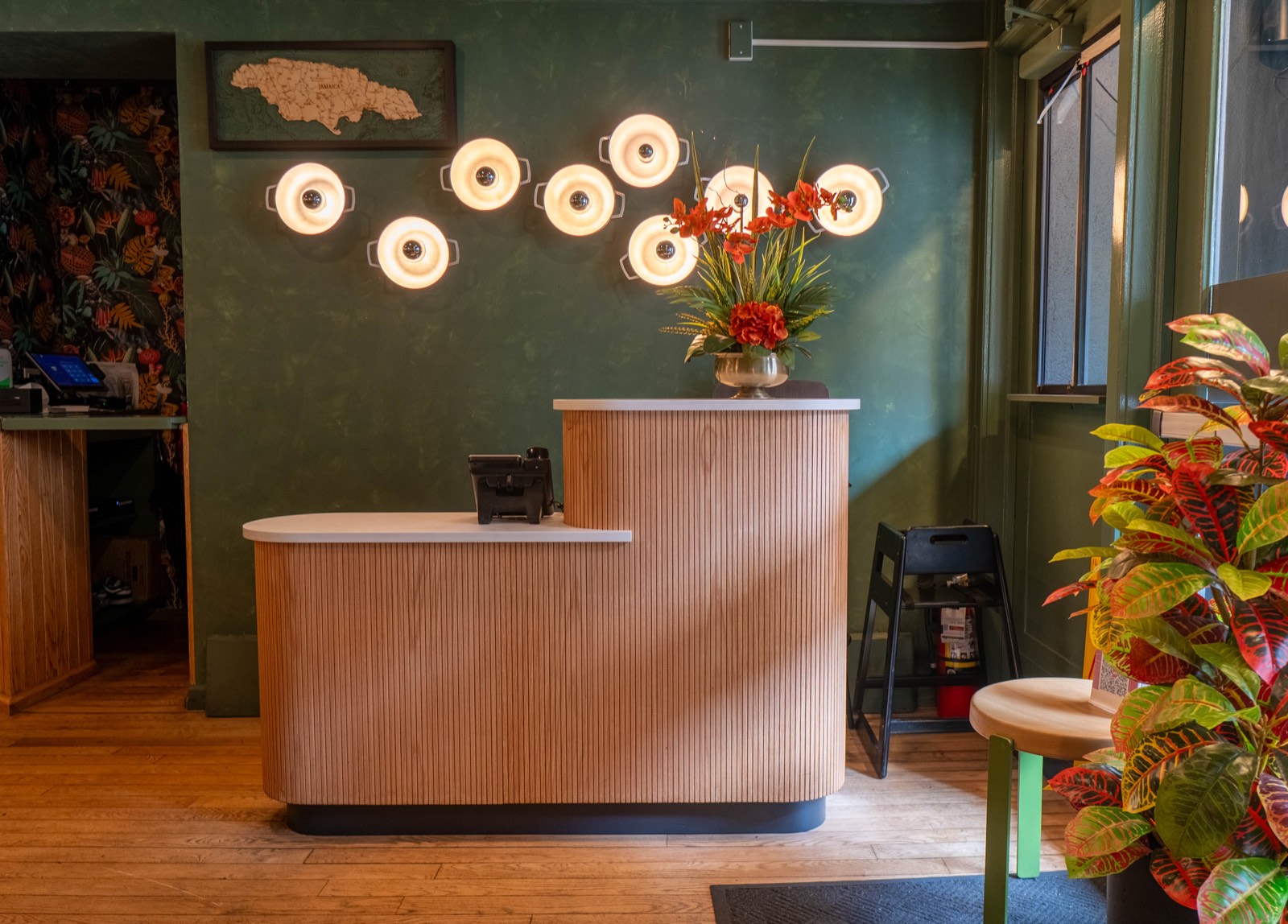 A two-tier host stand in fluted light oak with a pale stone top, against dark green plaster hung with a cluster of ring lights and a framed relief map of Jamaica