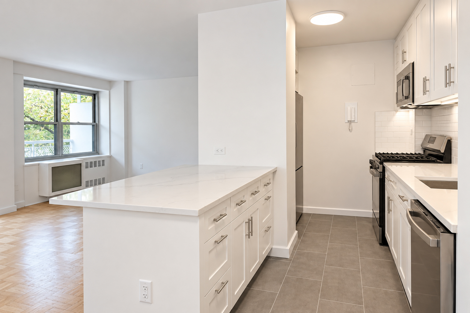 Galley kitchen with a peninsula of drawers and a stone top, opening to a living room with parquet floor and a window onto trees