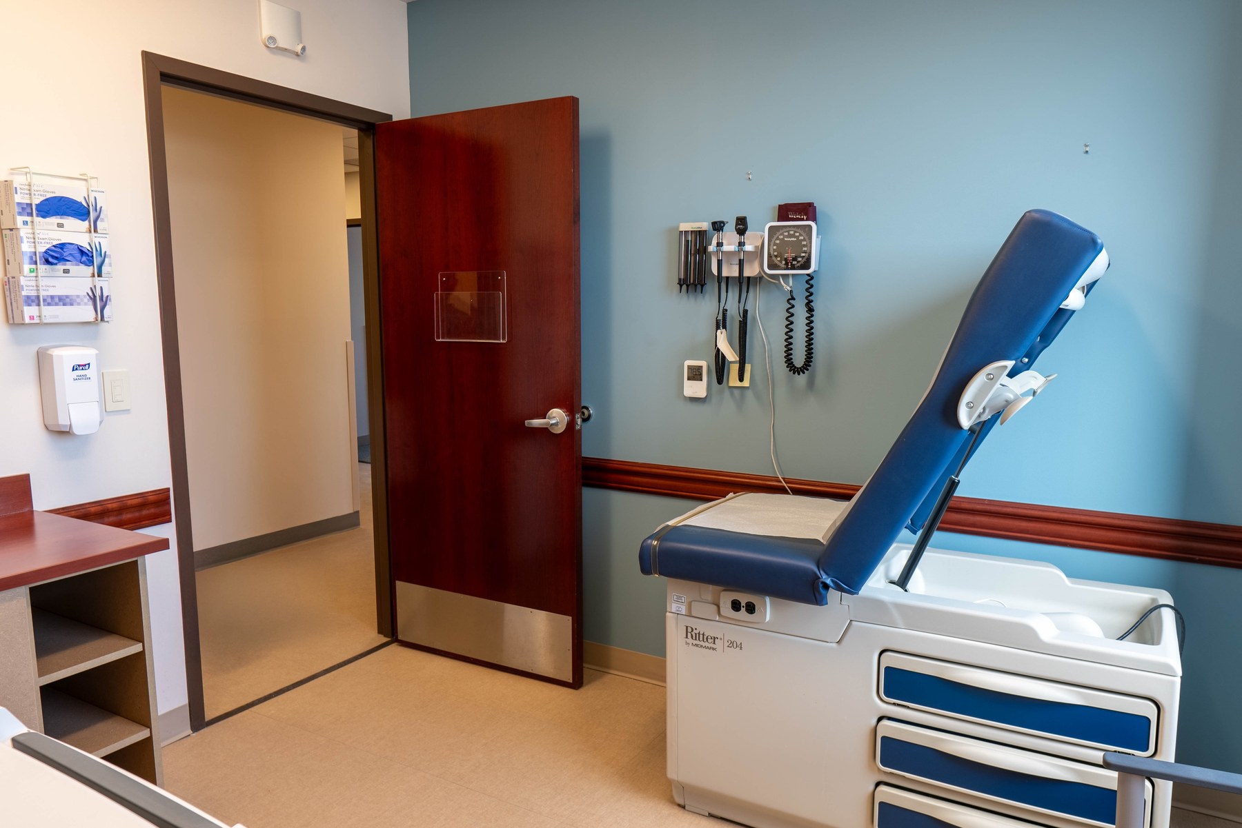 An exam table against a blue wall, with a wall-mounted diagnostic set and blood pressure gauge above it, a timber chair rail, and a solid-core door with a stainless kick plate