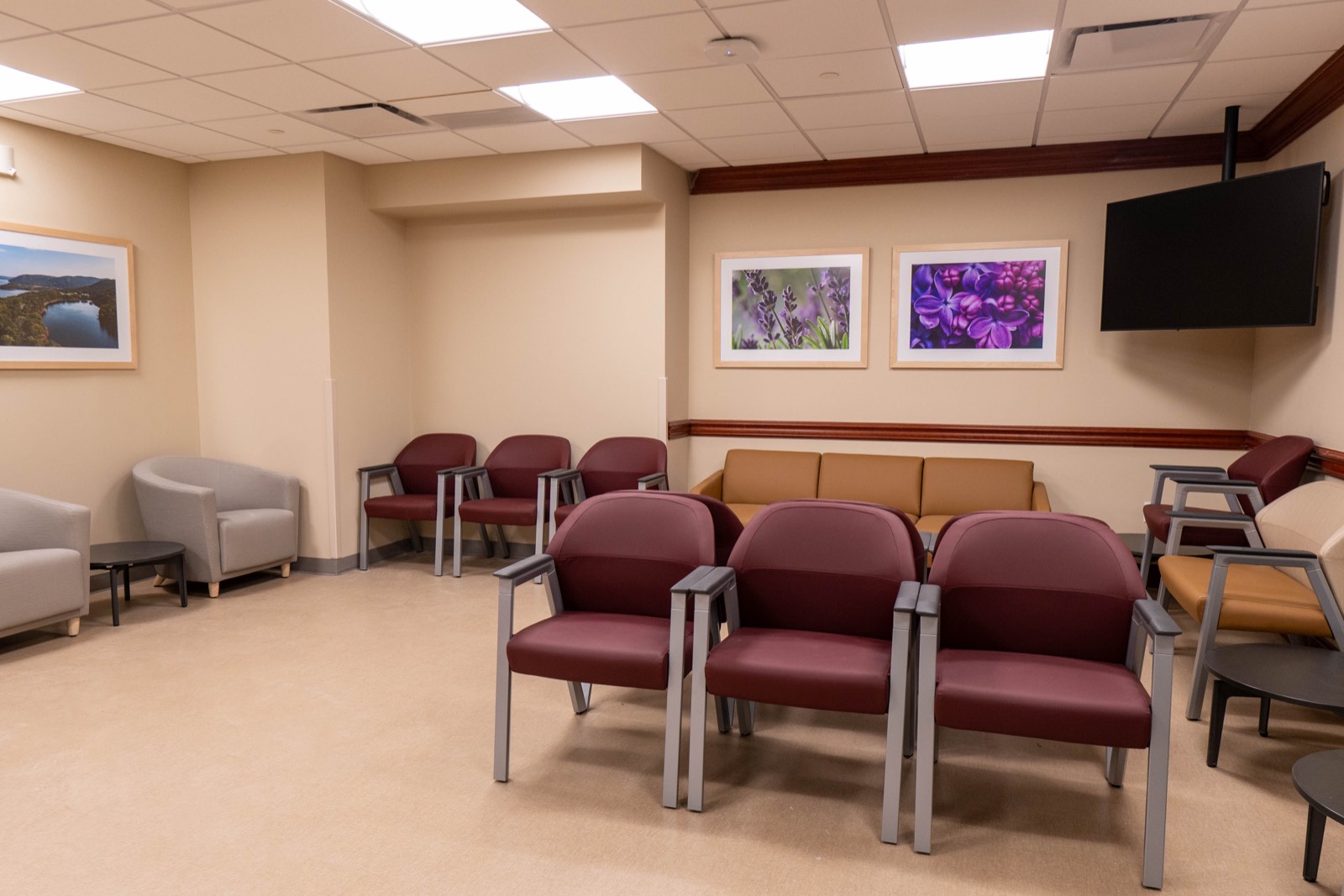 Rows of wipe-clean upholstered armchairs on metal frames, a timber chair rail running the perimeter, recessed troffers in a lay-in ceiling, and a wall-mounted screen in the corner