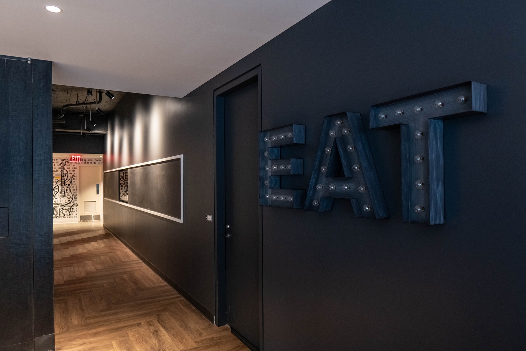 Marquee letters spelling EAT in stained blue timber with exposed bulbs, mounted on a dark corridor wall beside a framed chalkboard panel, above a herringbone wood floor
